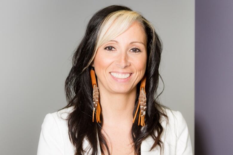 A woman with black hair with a blonde streak has a wide smile. She is wearing long feather earrings. 