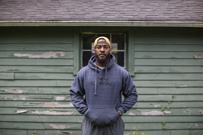 A Black man is pictured in a blue hoodie and yellow baseball cap in front of a green building.