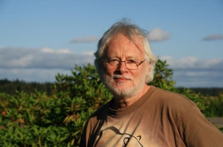 A man with grey hair and a grey beard wears T-shirt in front of foliage and blue skies. 