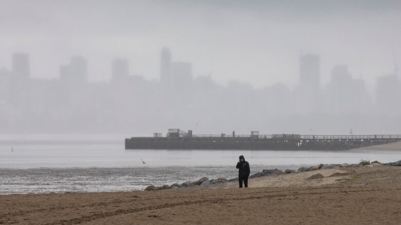 A person wearing a rain jacket walks on a cloudy, rainy beach in Vancouver with the city skyline obscured by fog in the background.