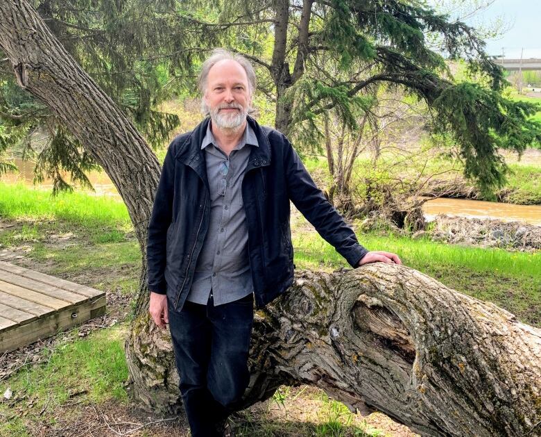 A man in a light jacket and blue shirt stands in the outdoors, with one hand on a tree.