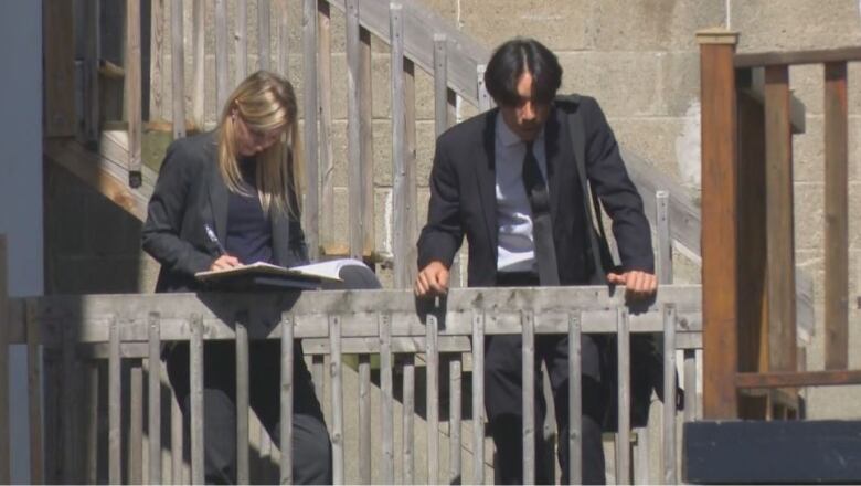 A woman, left, and a man, right, both wearing black suits, leaning on a wooden railing. The woman is writing in a book.