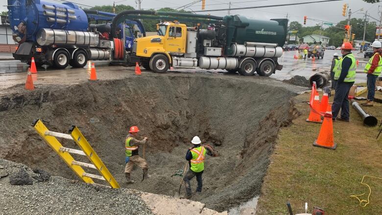 Workers in high-vis vests stand with shovels in a large hole on a street with trucks above them