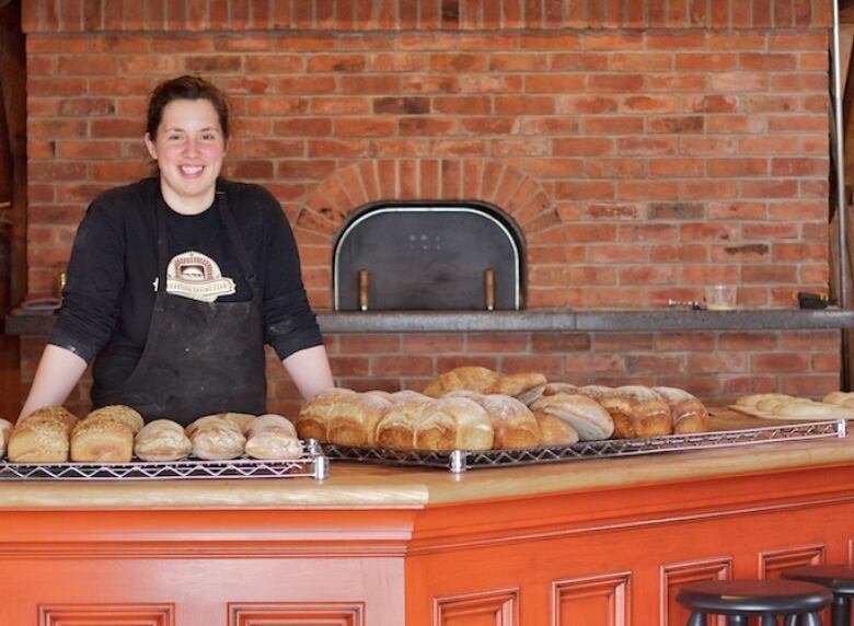 A chef wearing a black shirt and apron standing behind loaves of bread. 