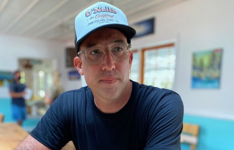 A man in a blue shirt, light blue hat and glasses sits at a table in a small restaurant.