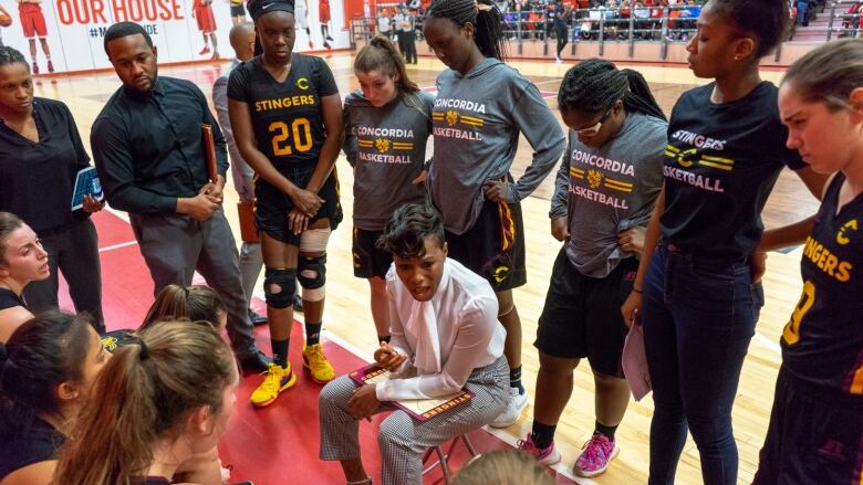 A woman in pants, a white blouse and heels sits in the centre of a huddle of female players on the edge of a basketball court, giving them instructions.