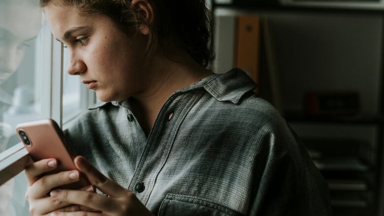 A young person holds a phone and looks out a window.