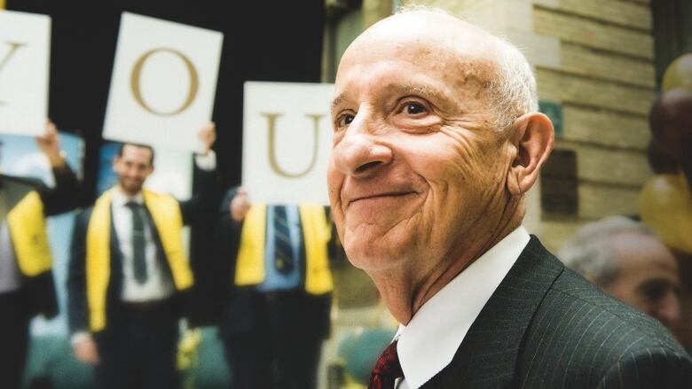 An older, balding man in a suit smiles with his head slightly turned over his left shoulder