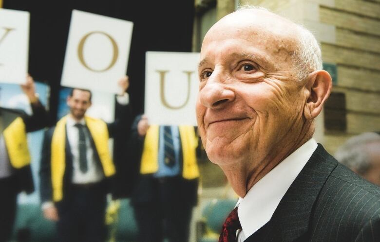 An older, balding man in a suit smiles with his head slightly turned over his left shoulder