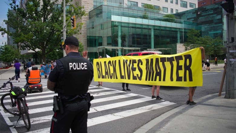 A police officer stands on a sidewalk. Behind him, people hold a Black Lives Matter banner.