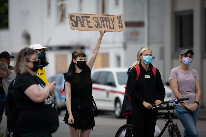 A person wearing a facemask holds up a sign that reads 'Safe Supply Now' at a rally.