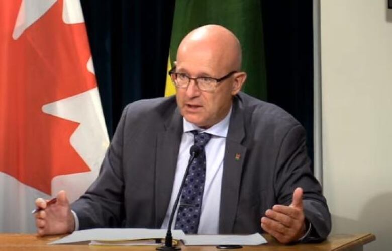 Warren Kaeding is sitting at a wood table with his hands out and a canadian flag behind him