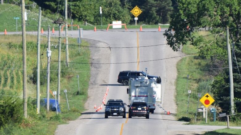 Police vehicles down roadway that's been closed off to traffic. It's woods and farmland beside the road.