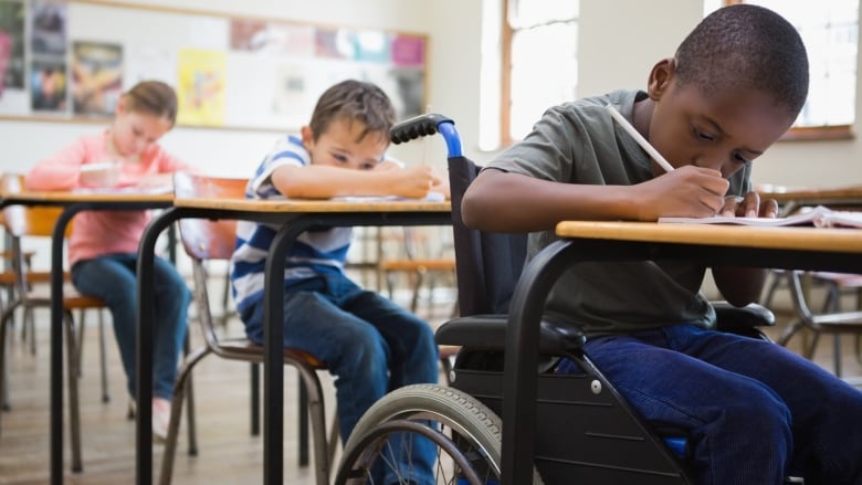 Three young students are shown writing at desk. The boy closest to the front is in a wheelchair.