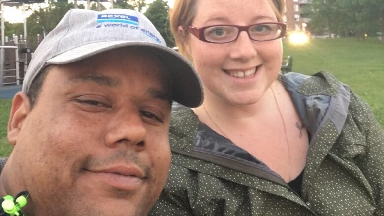 A Black man wearing a hat is seen taking a selfie with a white woman wearing glasses.