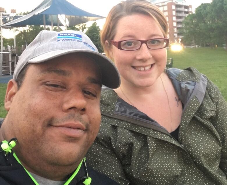 A Black man wearing a hat is seen taking a selfie with a white woman wearing glasses.