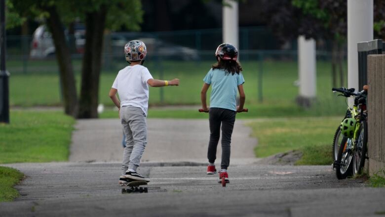 One child on a scooter and another child on a skateboard play in a park.