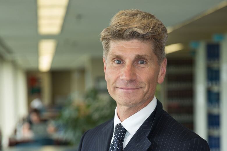 A middle aged man with blonde hair who is wearing a black suit and tie poses for a photo. 