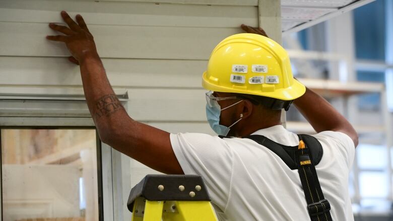 A construction worker with a yellow hard hat has their hands pressing against the side of a home.