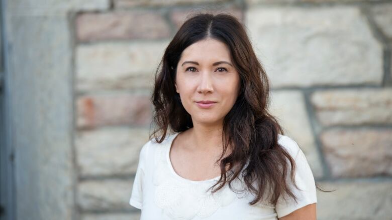 A woman with long hair wears a white shirt and stands in front of a stone wall.