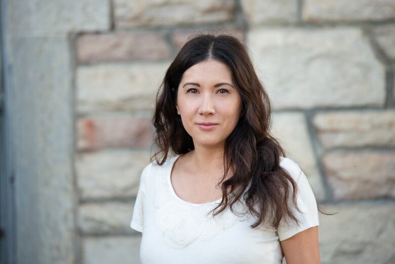 A woman with long hair wears a white shirt and stands in front of a stone wall.