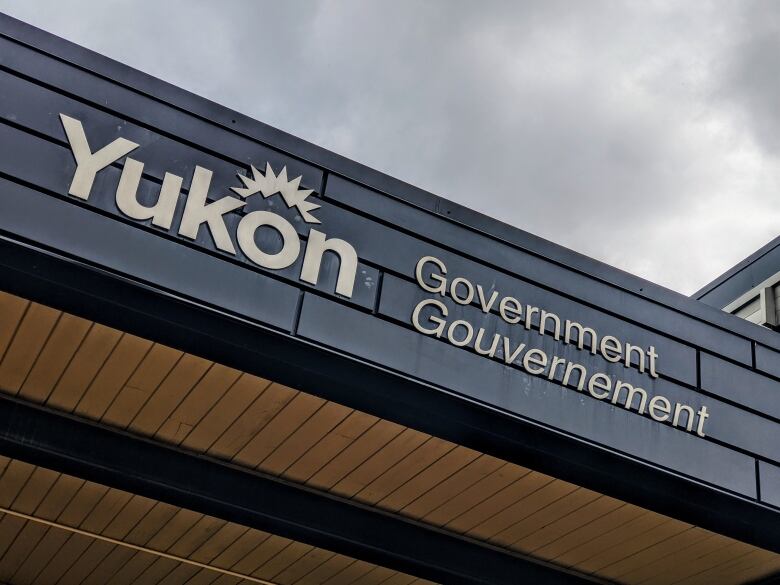 A building facade with the words Yukon government in the forefront of a grey sky.