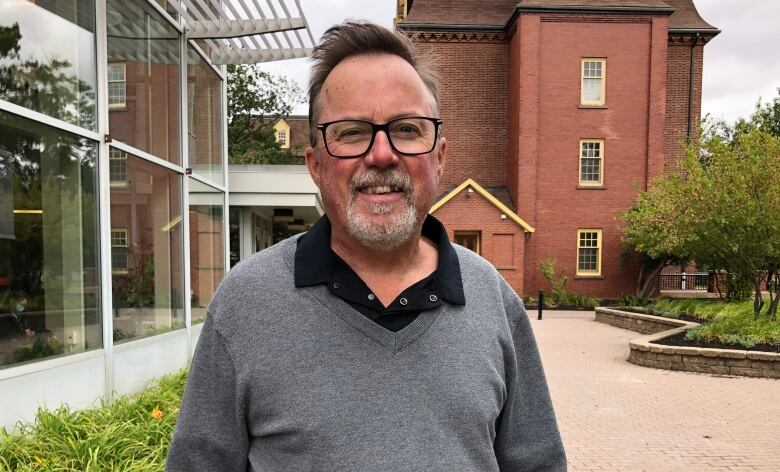 A man with glasses a beard and a blue sweater vest stands on a university campus