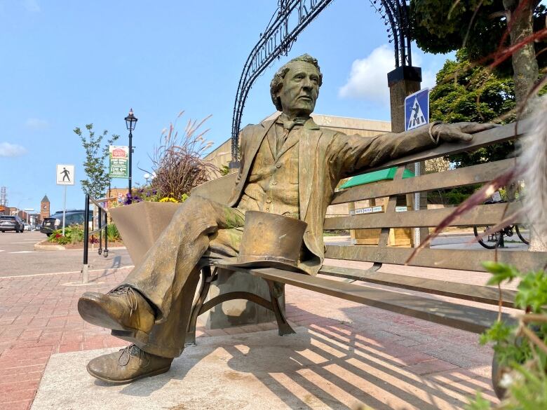 A bronze statue of a man in 19th-century clothing sitting on a park bench is seen on a city street with the Confederation Centre of the Arts in the background. 