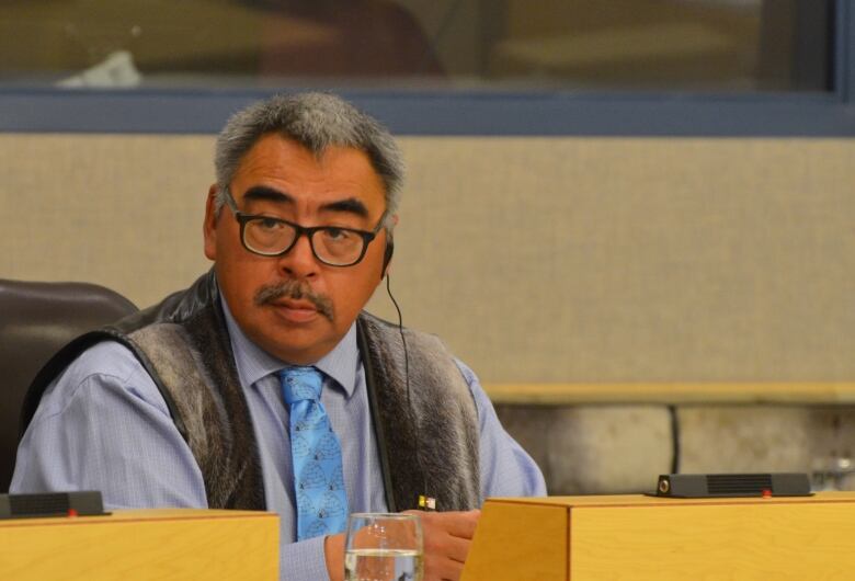 An Inuk man with salt and pepper hair sits in the Nunavut Legislature wearing a sealskin vest, matching his haircolour.