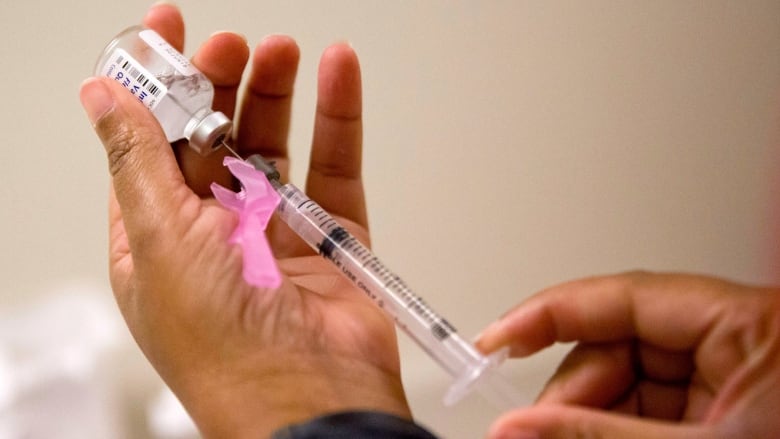 A hand holding a syringe draws vaccine from a vial held in their other hand.