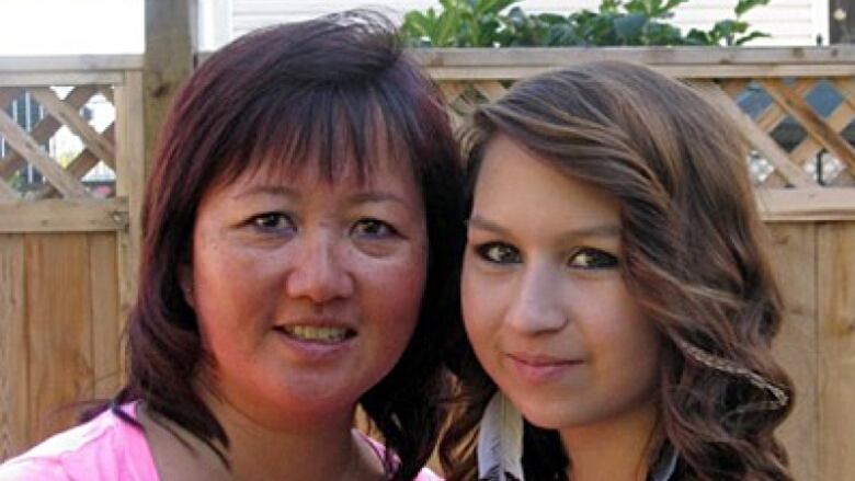 A middle-aged woman with black hair stands next to a teenage girl with long brown hair and feather earrings. Their heads are close together and they are smiling while standing in a yard with a lattice fence in the background.