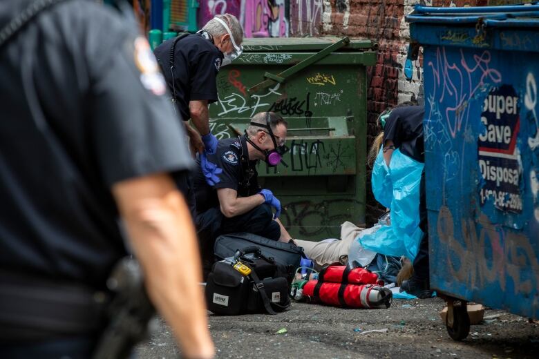 Paramedics attend a person suspected of having a drug overdose beside a dumpster in an alley.