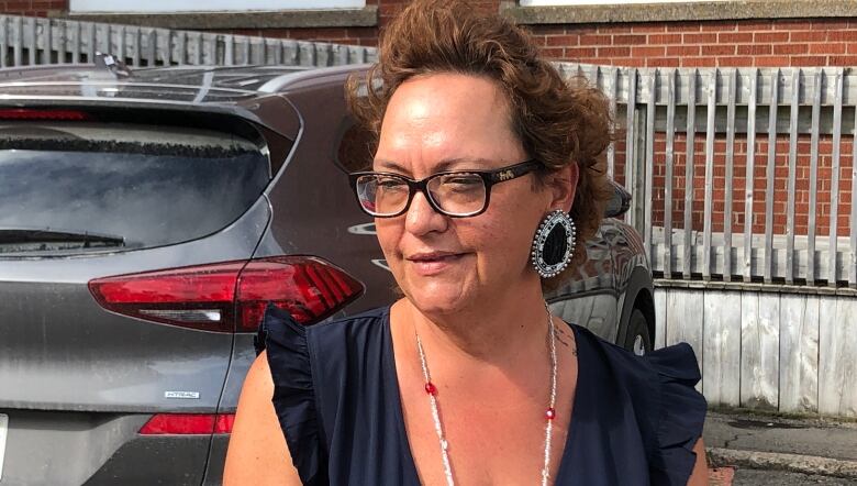 A woman wearing glasses and a large necklace speaks to the camera.