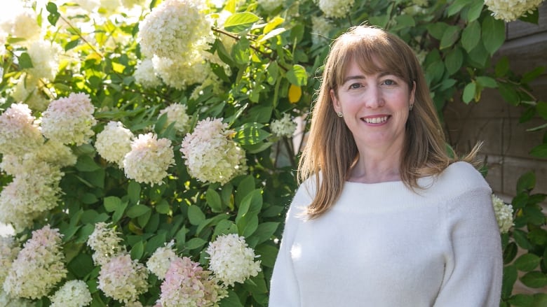 A woman in a white sweater with long blonde hair smiles for the camera in front of a flower bush with large white blossoms.