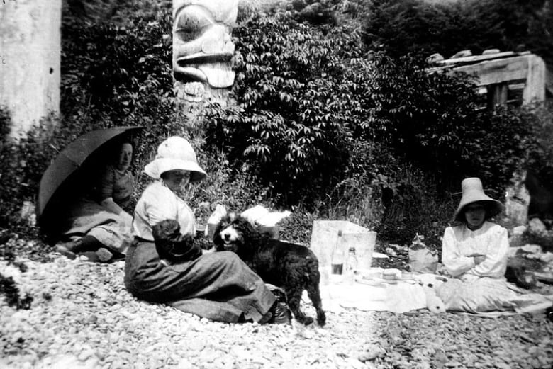 A black and white photo from the late 19th century or early 20th century showing a woman in long dress and hat mostly obscuring her face sitting on the ground with a dog. Two other people sitting, one holding an umbrella, and a totem pole are in the background.