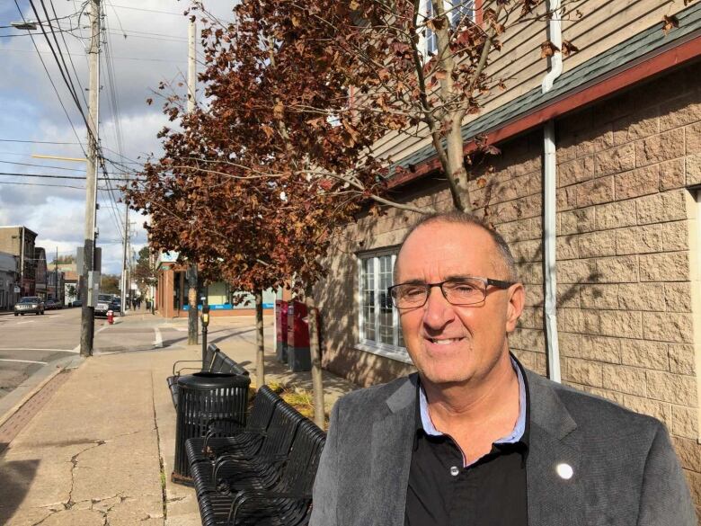 A man in a grey suit jacket and glasses stands on a downtown sidewalk.