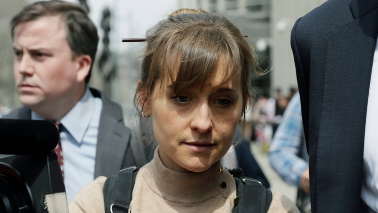 A close-up photo of woman with her hair in a pony tail walks outside next to a man in a suit with another man in a suit following close behind. 