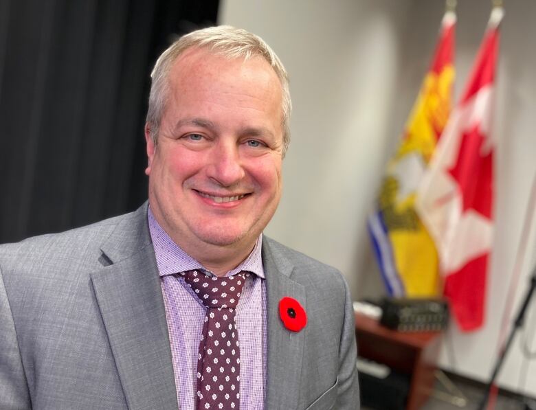 A man in a suit smiles at the camera, with the Canadian and New Brunswick flags in the foreground.