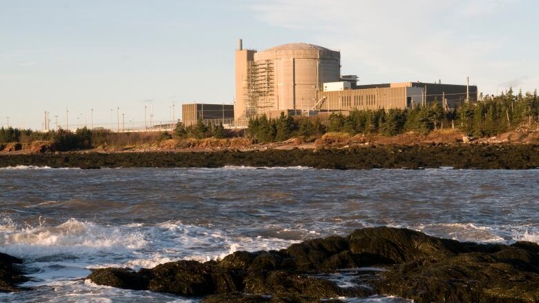 A concrete building with a squat, round tower sits on the edge of land with rocks and water in the foreground.