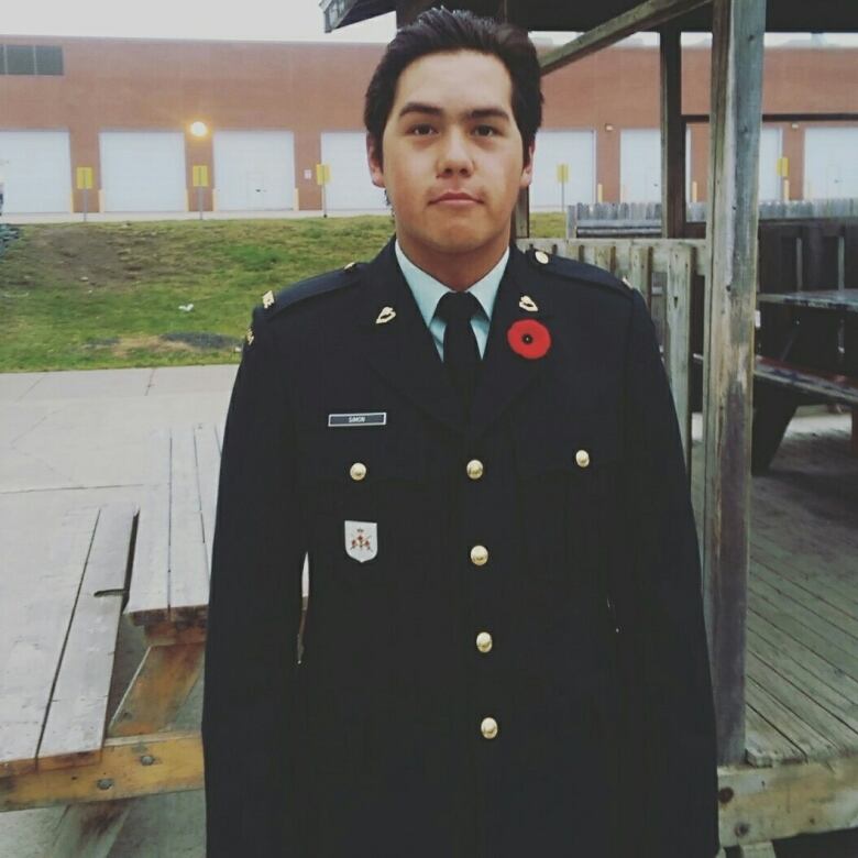 A young man with short dark hair combed back wearing a dark-coloured suit with Canadian military emblems and a poppy on his left lapel. He is standing outdoors in front of a wooden picnic table, with a brick commercial building in the distant background.