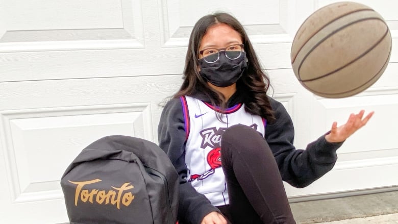 A teenaged girl with long black hair and glasses, and wearing a face mask, sits on a gymnasium floor, tossing a basketball with one hand.