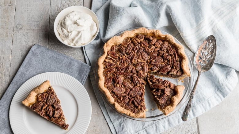 Overhead shot of a pecan pie in a glass pie plate, sitting on a light blue kitchen towel, on a grey wooden surface. A piece of the pie sits on a white plate in the bottom left corner.