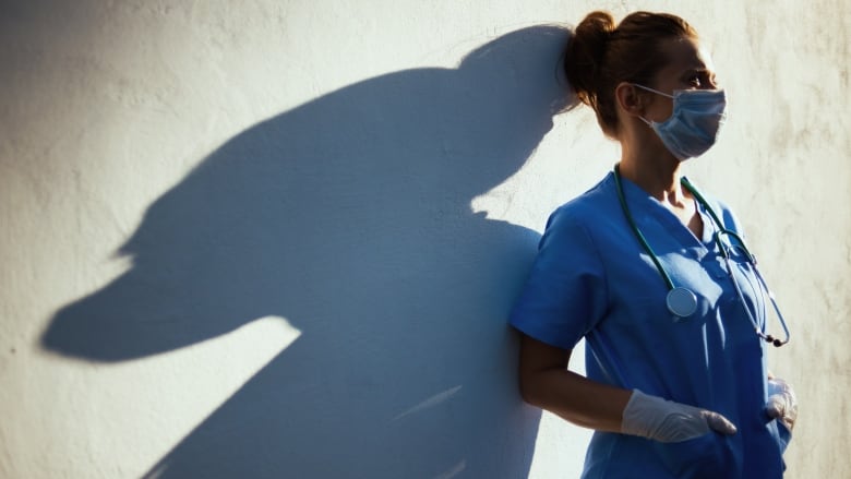 A tired female doctor in scrubs with stethoscope, medical mask and rubber gloves stands outdoors during the coronavirus pandemic.