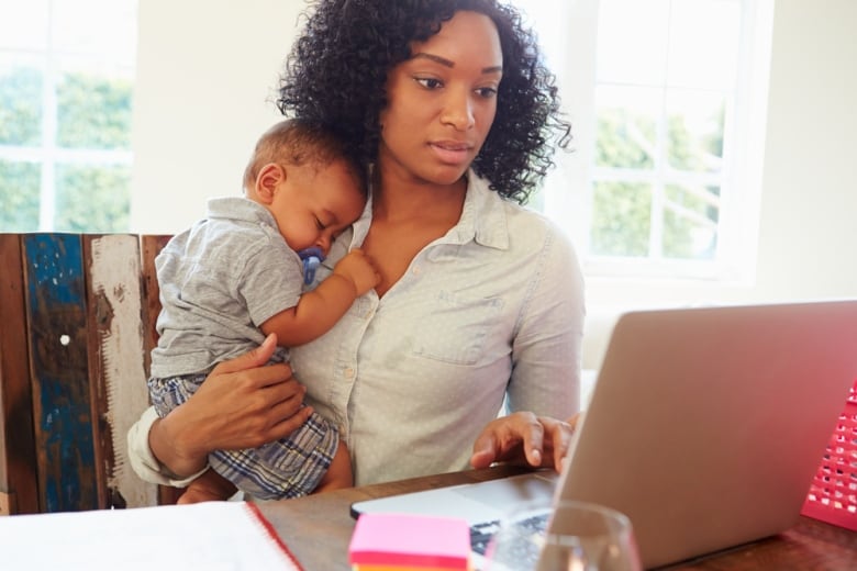 A woman holds a child while sitting at a desk and working at a laptop computer. 