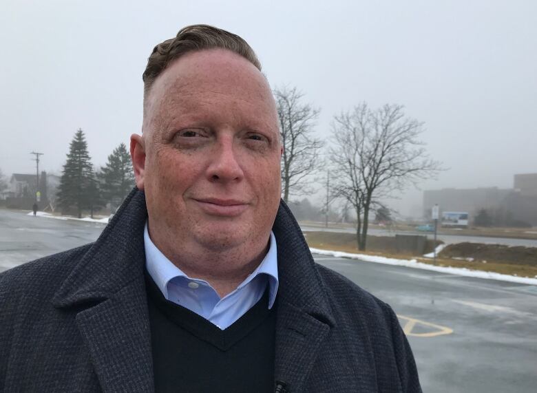 Man in dress shirt and jacket standing outdoors with parking lot behind him.