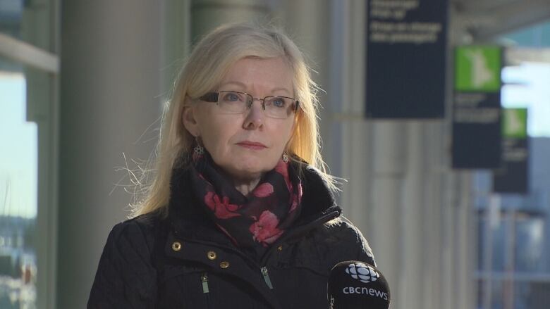 A white woman with glasses and blonde hair stands outside an airport terminal wearing a coat and scarf