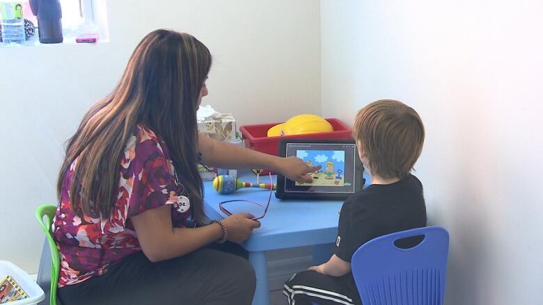 A woman and a boy seated at a small blue plastic table look at a tablet.