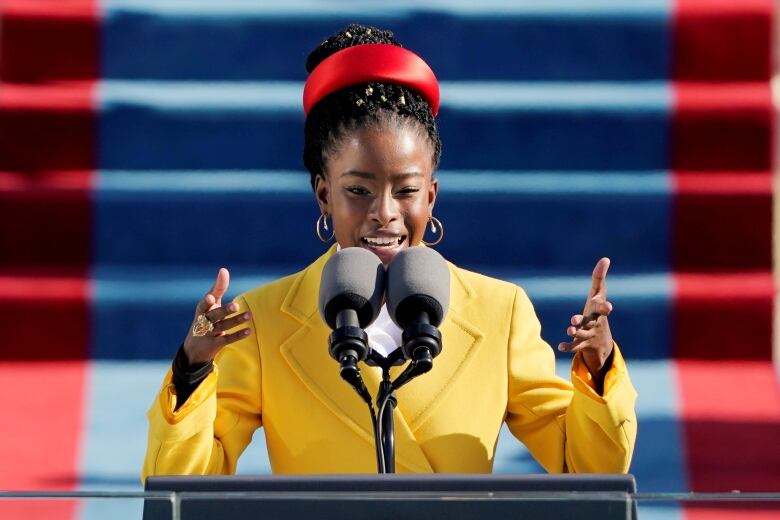 A woman in a bright yellow coat and red hat destrures and smiles as she speaks into a microphone