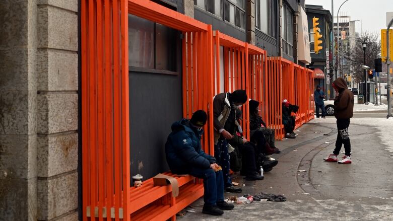 People sit on bright orange benches along the wall of a downtown building.