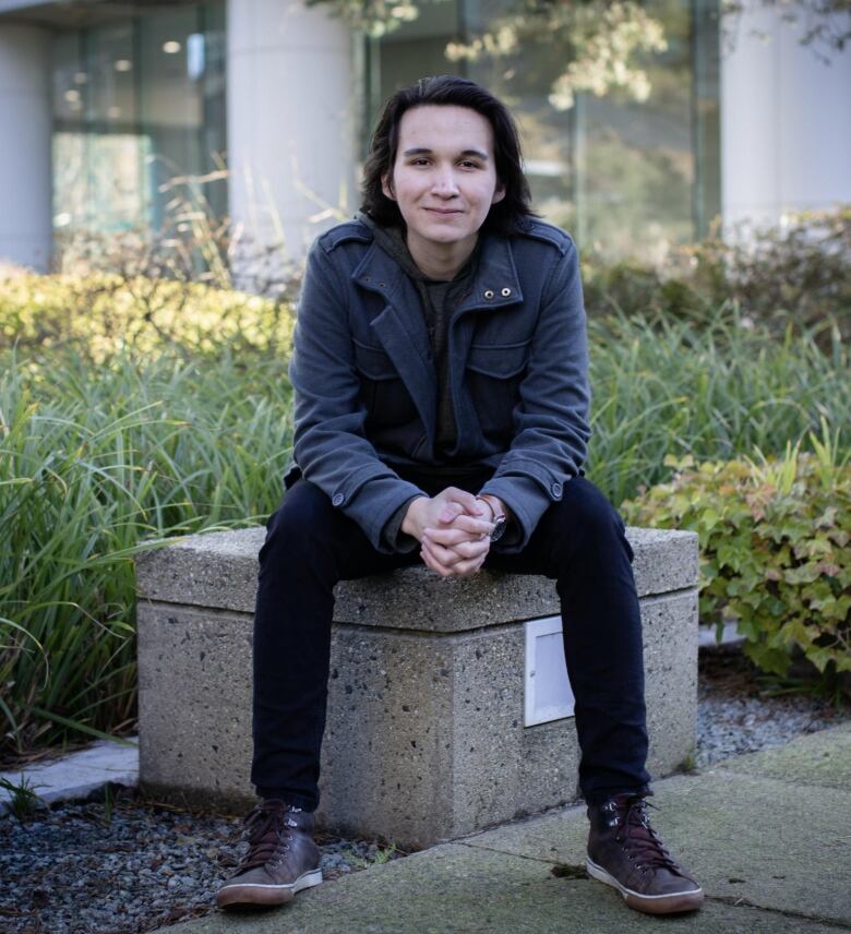 A man with medium-length black hair sits on a small bench.